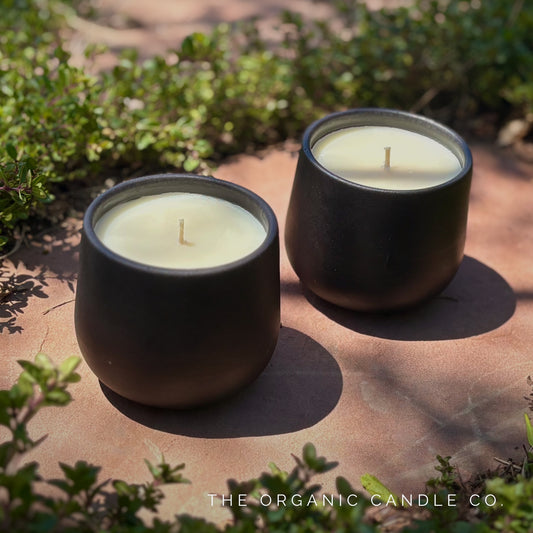 Two organic candles in black ceramic containers on a stone surface with greenery around.