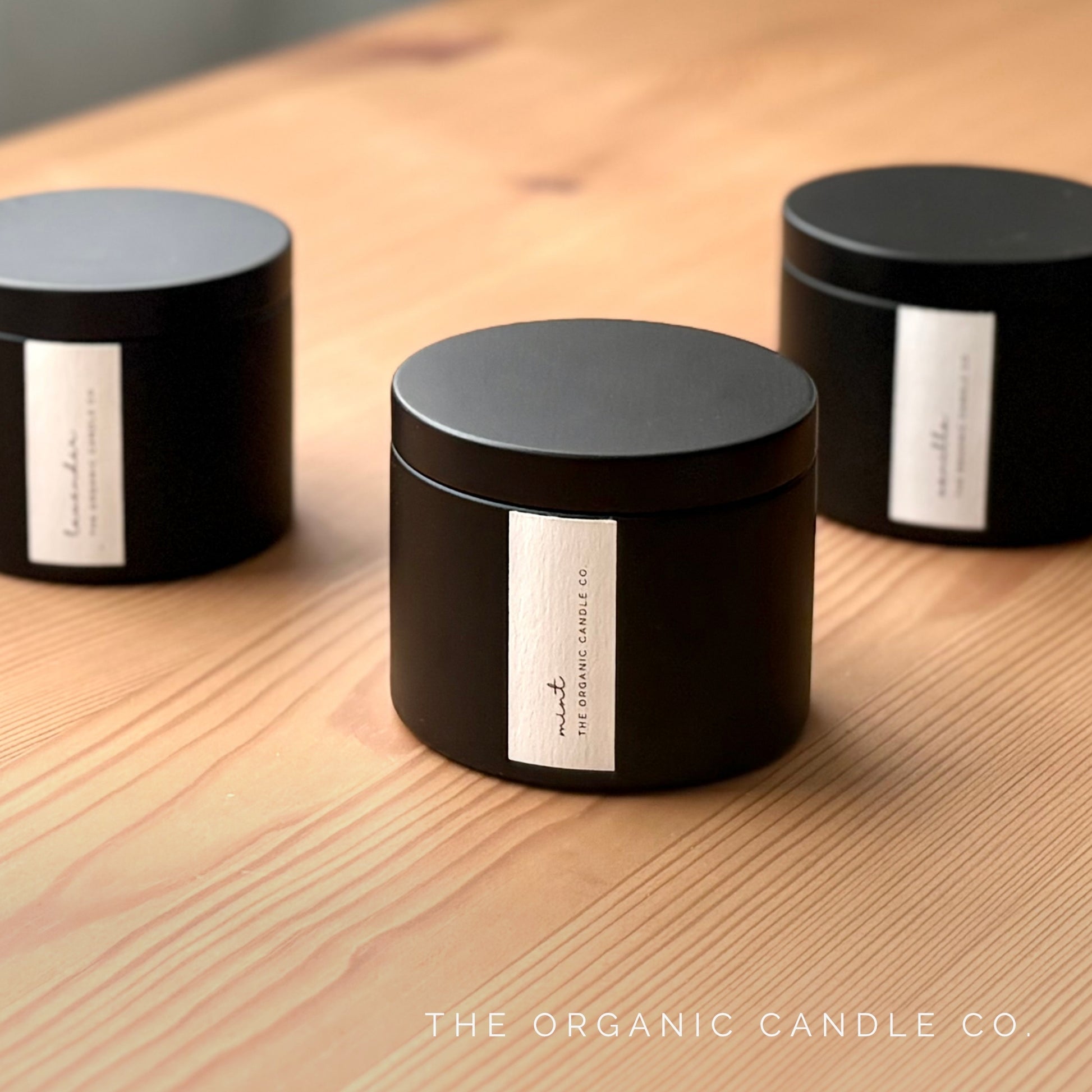 Three black candles with labels on a wooden surface, featuring 'The Organic Candle Co'.