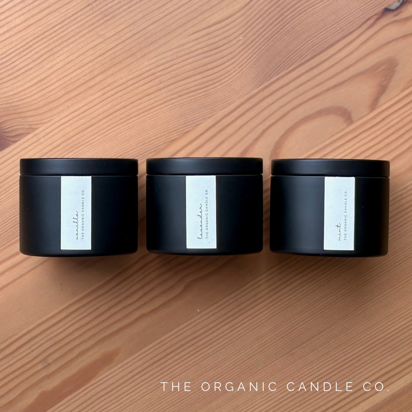 Three black candles with white labels on a wooden surface, branded 'The Organic Candle Co'.