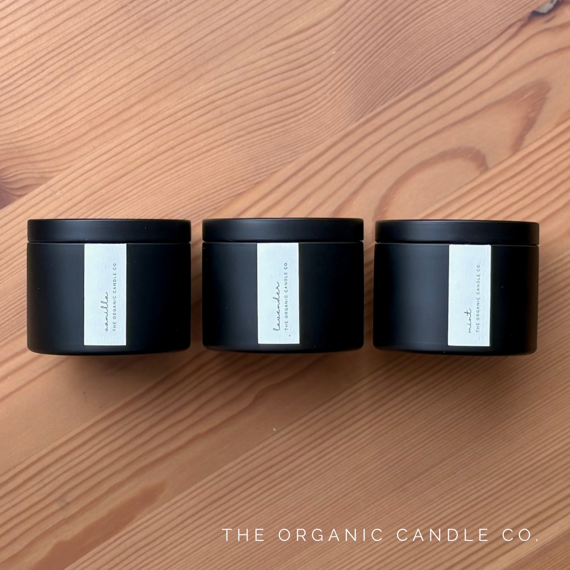 Three black candles with white labels on a wooden surface, branded 'The Organic Candle Co'.