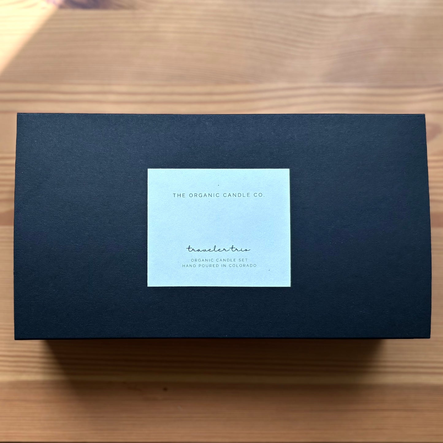 A black rectangular product box with a light blue-white square label in the center, placed on a light wood surface. The label reads 'The Organic Candle Co.'
