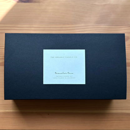 A black rectangular product box with a light blue-white square label in the center, placed on a light wood surface. The label reads 'The Organic Candle Co.'