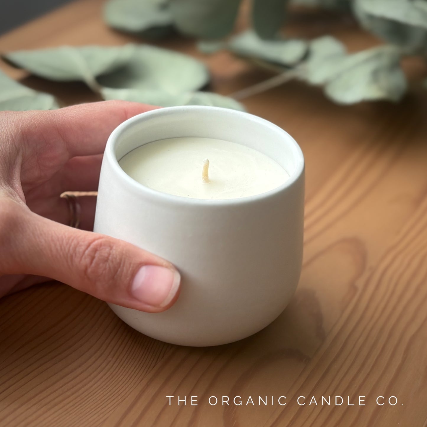 Hand holding a white organic candle on a wooden surface with 'The Organic Candle Co.' branding.