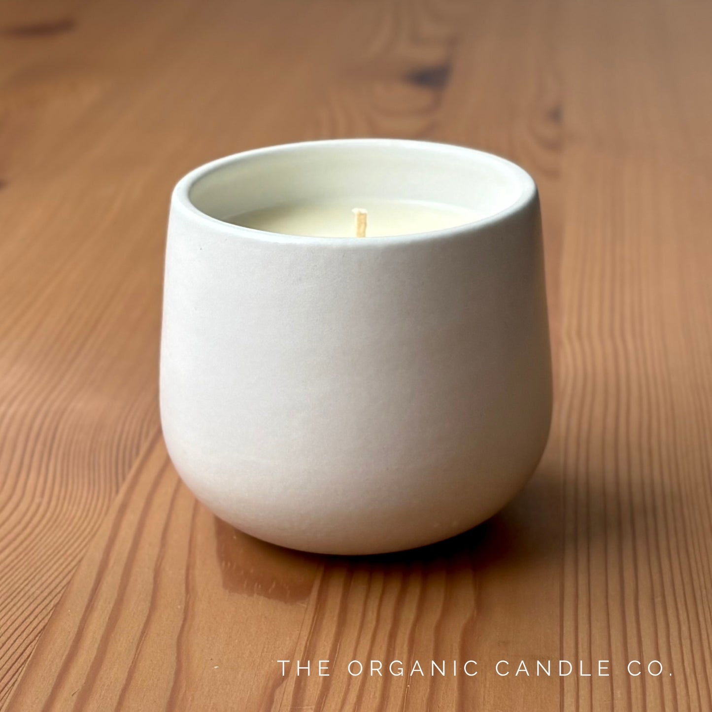 White organic candle in a ceramic holder on a wooden surface with 'The Organic Candle Co.' branding.