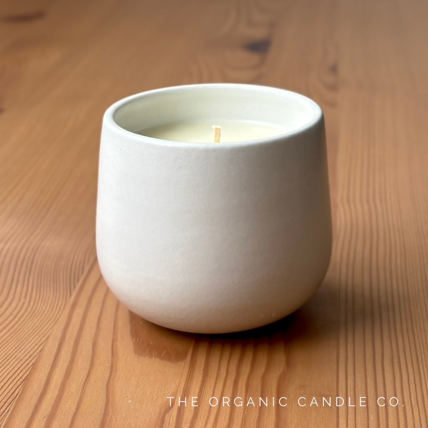 White organic candle in a ceramic vessel on a wooden surface with 'The Organic Candle Co.' branding.