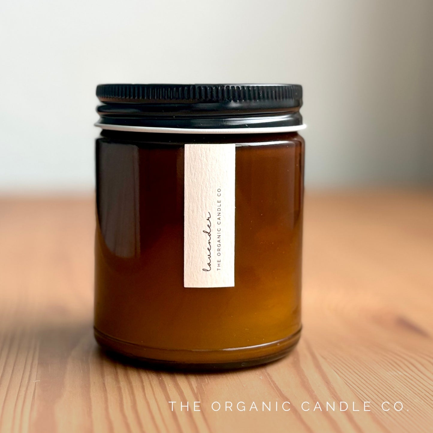 Organic candle in an amber glass vessel with black lid on a wooden surface, featuring 'The Organic Candle Co' label.
