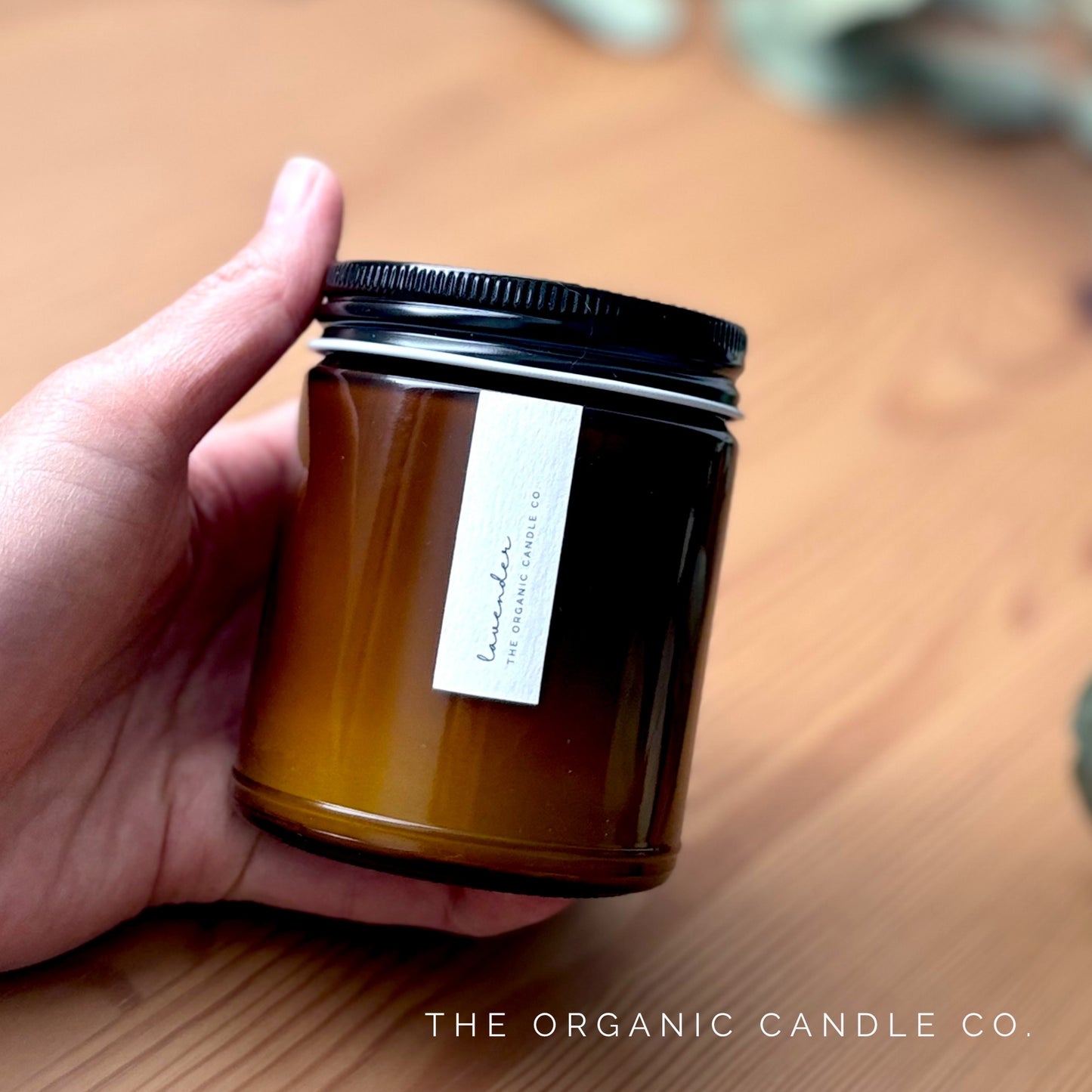 Hand holding an organic candle in an amber glass vessel with a black lid on a wooden surface, featuring 'The Organic Candle Co.'