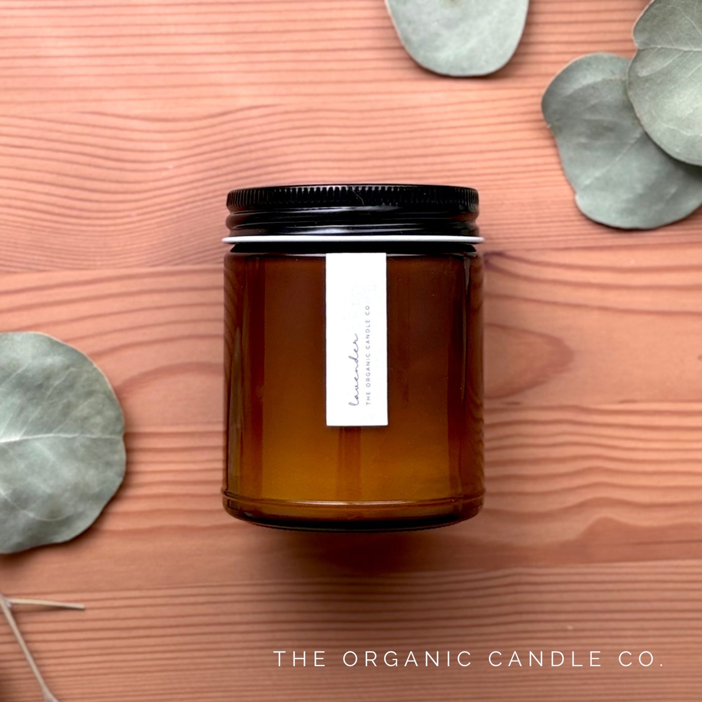 Organic candle in an amber glass vessel with black lid on a wooden surface with eucalyptus leaves.