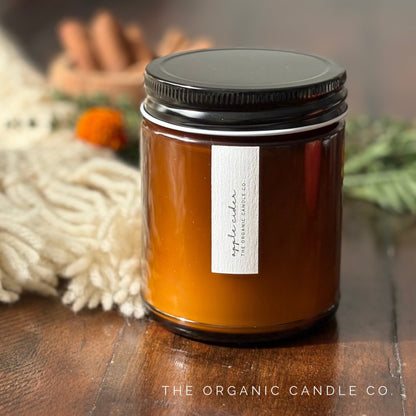 A candle in an amber glass jar labeled 'apple cider' by The Organic Candle Co., styled on a wooden surface with a cream fringed textile and cinnamon sticks in the background.