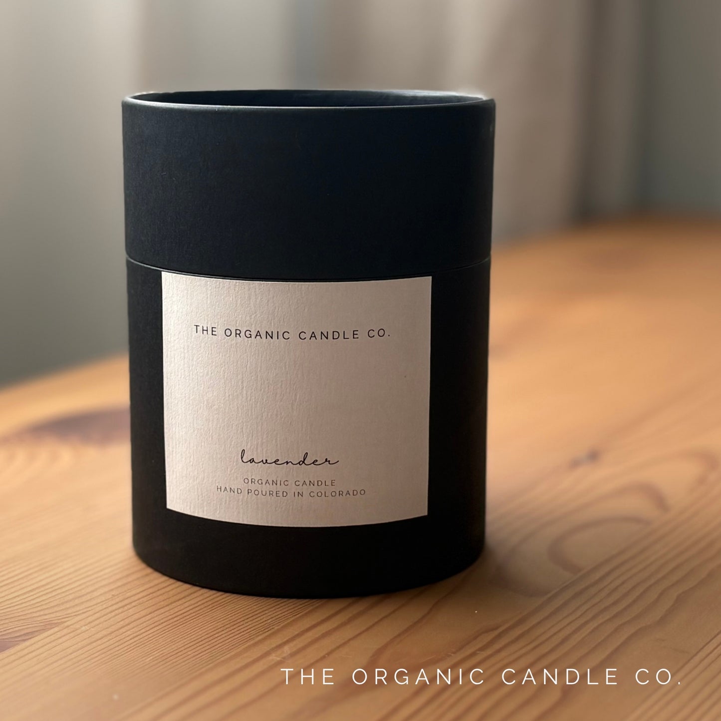 Black cylindrical container with a label on a wooden surface, featuring 'The Organic Candle Co.'