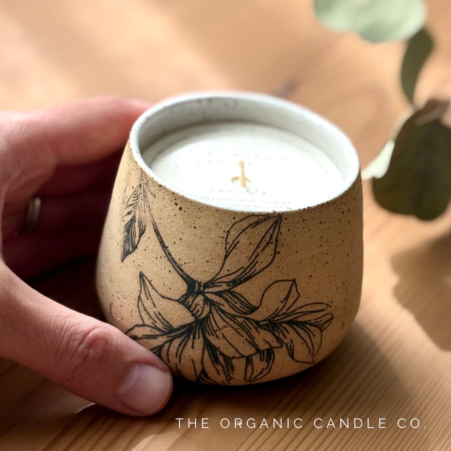 Organic candle in ceramic vessel with botanical design, held by a hand on a wooden surface.