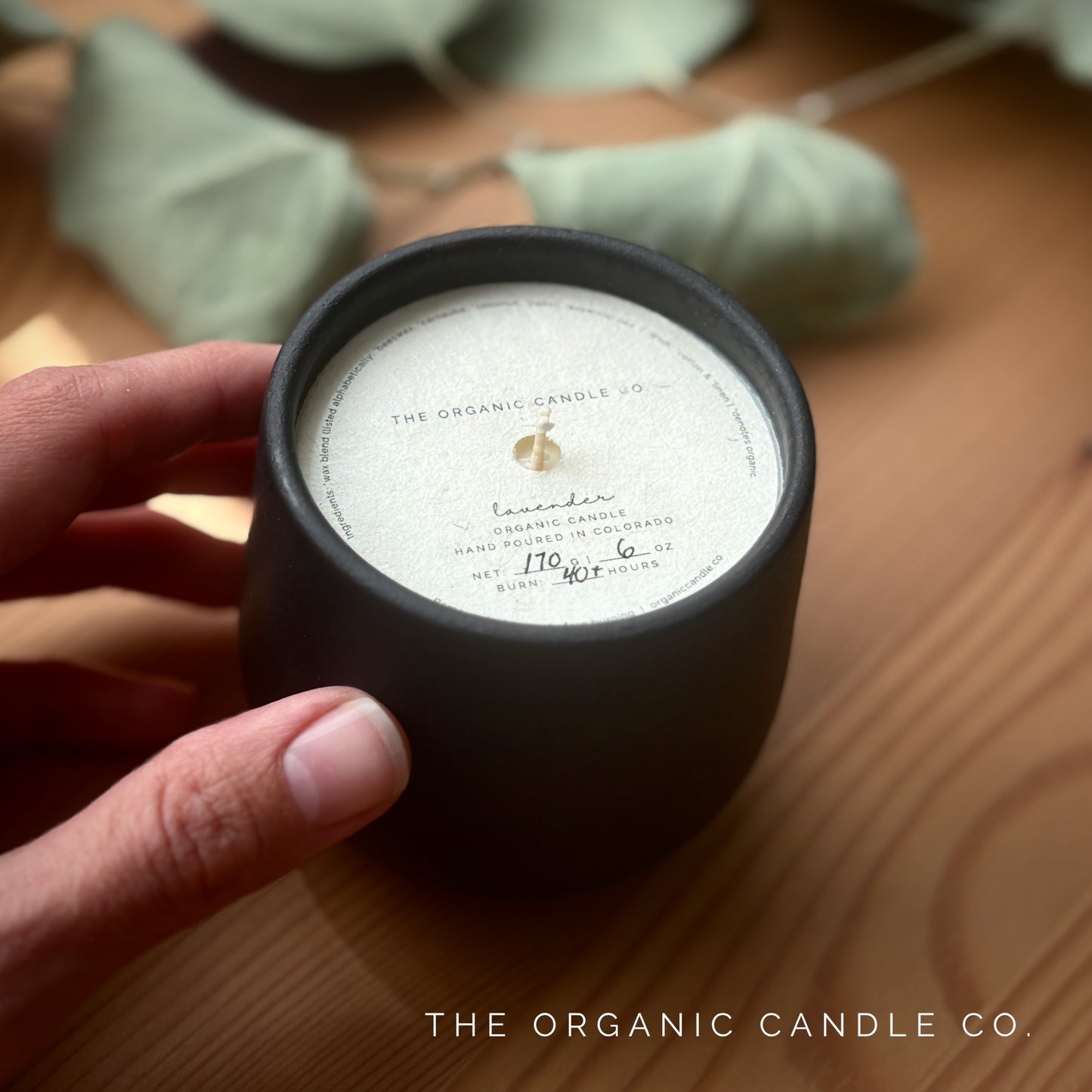 Organic candle in black ceramic vessel held by a hand on a wooden surface, featuring 'The Organic Candle Co.'