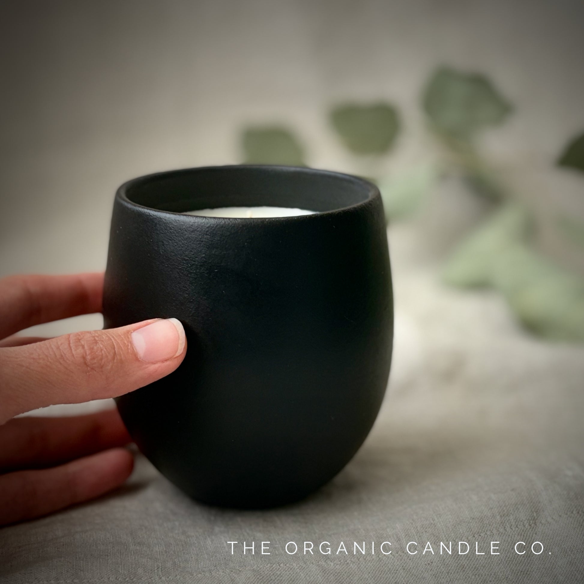 Black organic candle with a hand holding it, blurred greenery in the background, 'The Organic Candle Co.' branding.