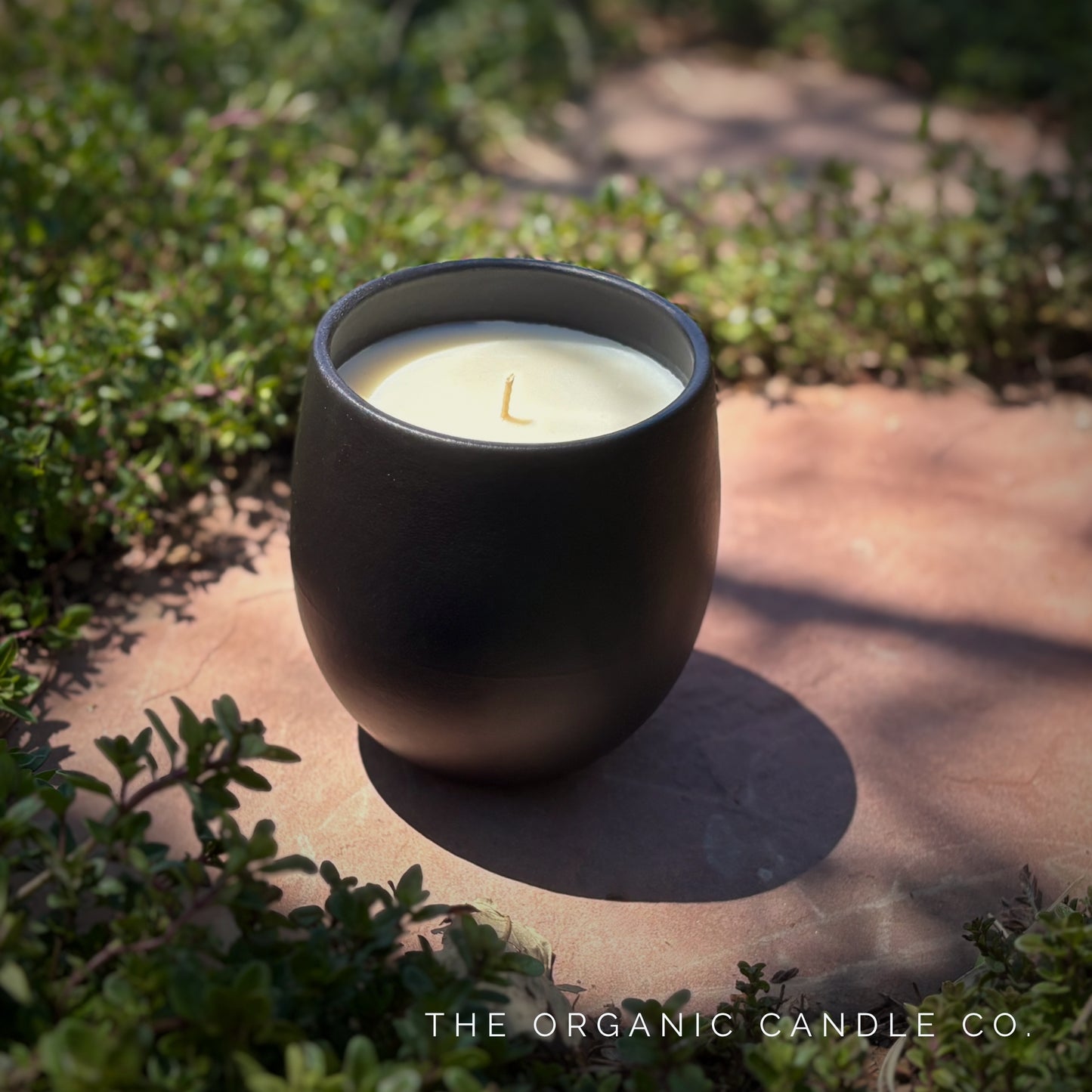 Organic candle in black ceramic vessel on a stone surface with greenery around