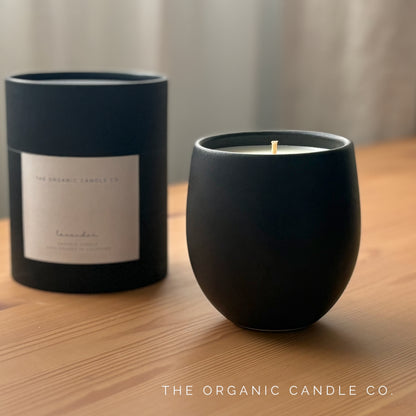 Organic candle in black ceramic vessel and black cylindrical packaging on a wooden surface, branded 'The Organic Candle Co'.