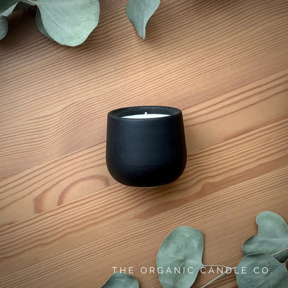 Organic candle in black ceramic vessel on a wooden surface with eucalyptus leaves, featuring 'The Organic Candle Co'.