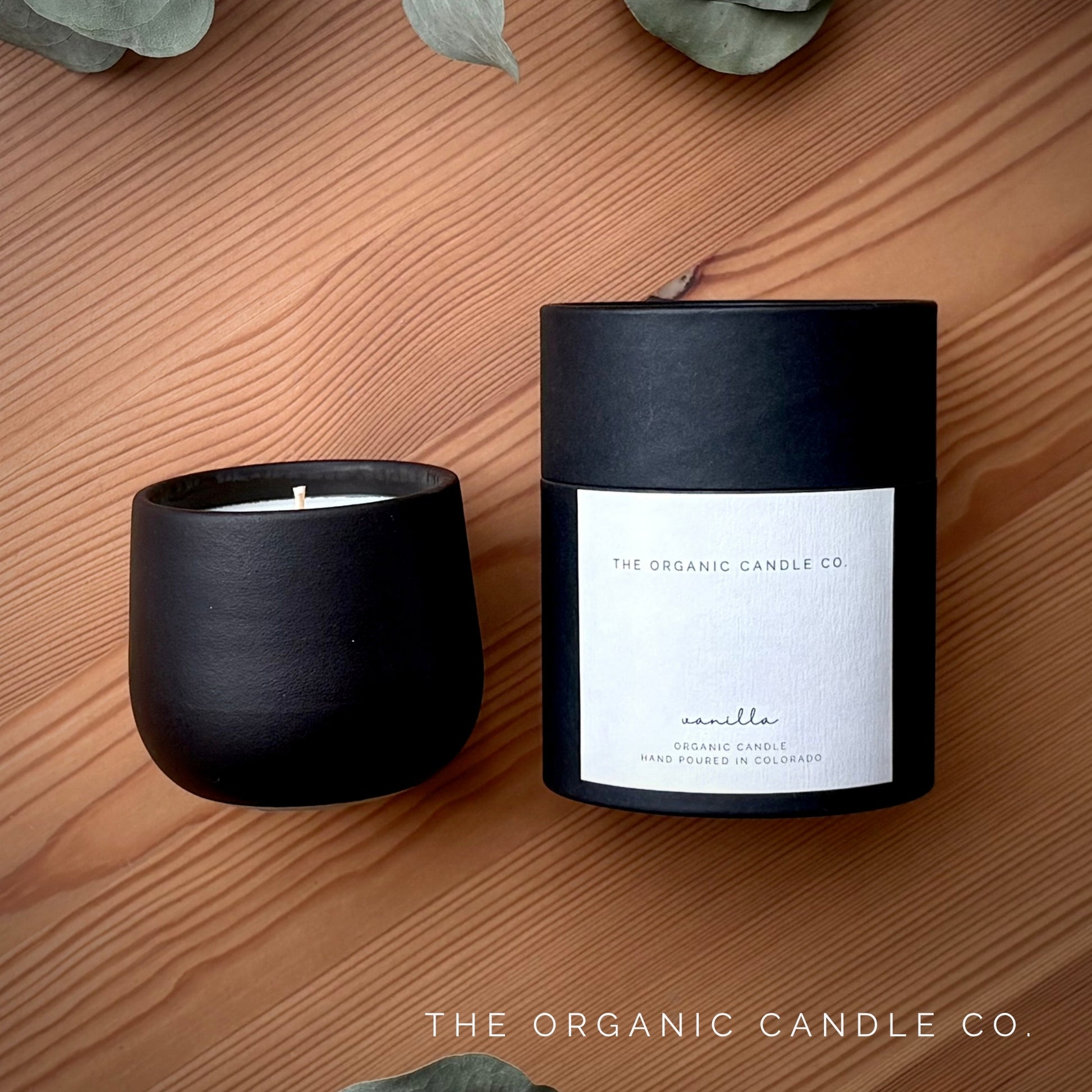 Organic candle in black ceramic vessel  from The Organic Candle Co. and its packaging on a wooden surface with green leaves.