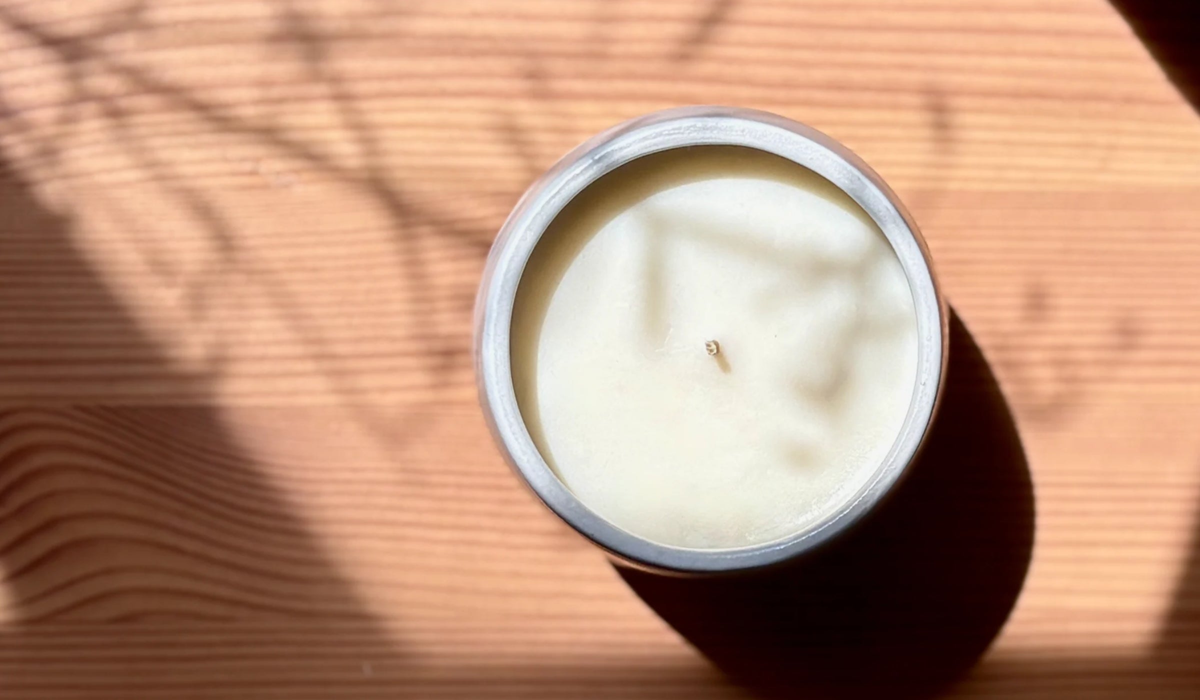 A top-down view of a white organic candle in a white ceramic, placed on a light wood grain surface with dappled sunlight and shadows.
