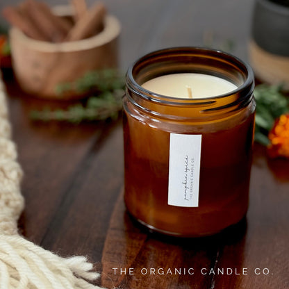 A candle in an amber glass jar labeled 'Pumpkin Spice' by The Organic Candle Co., styled on a wooden surface with a cream fringed textile and cinnamon sticks in the background.